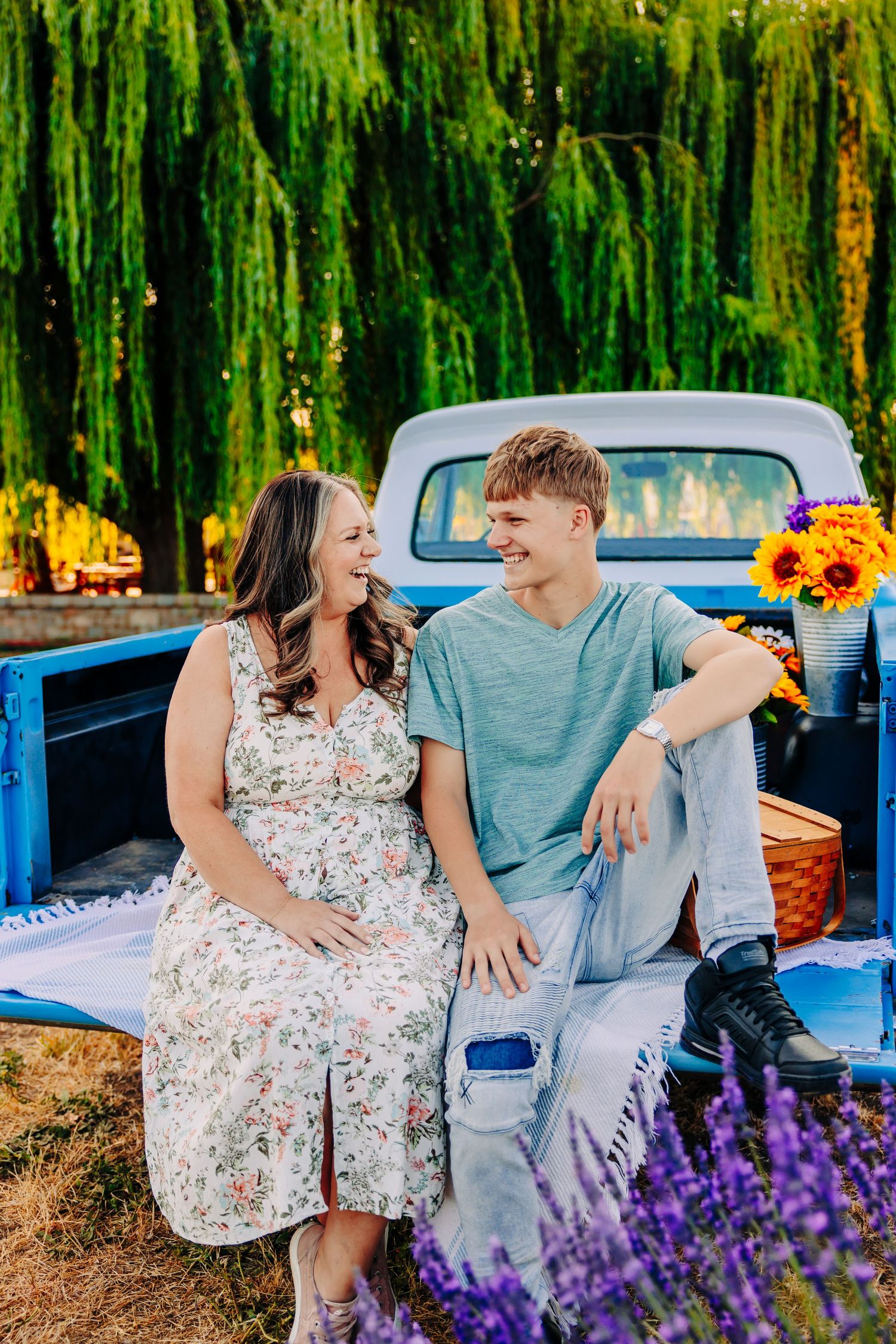 A romantic couple poses with a vintage blue pickup truck surrounded by vibrant sunflowers and weeping willows.