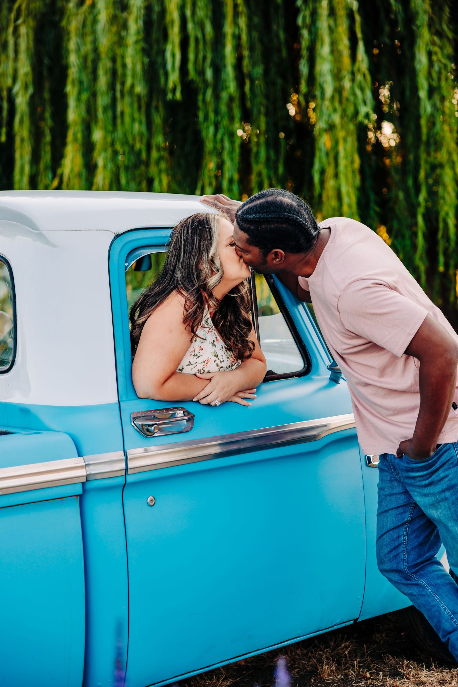 A couple shares a romantic kiss through the window of a classic light blue vintage car in front of willow trees.