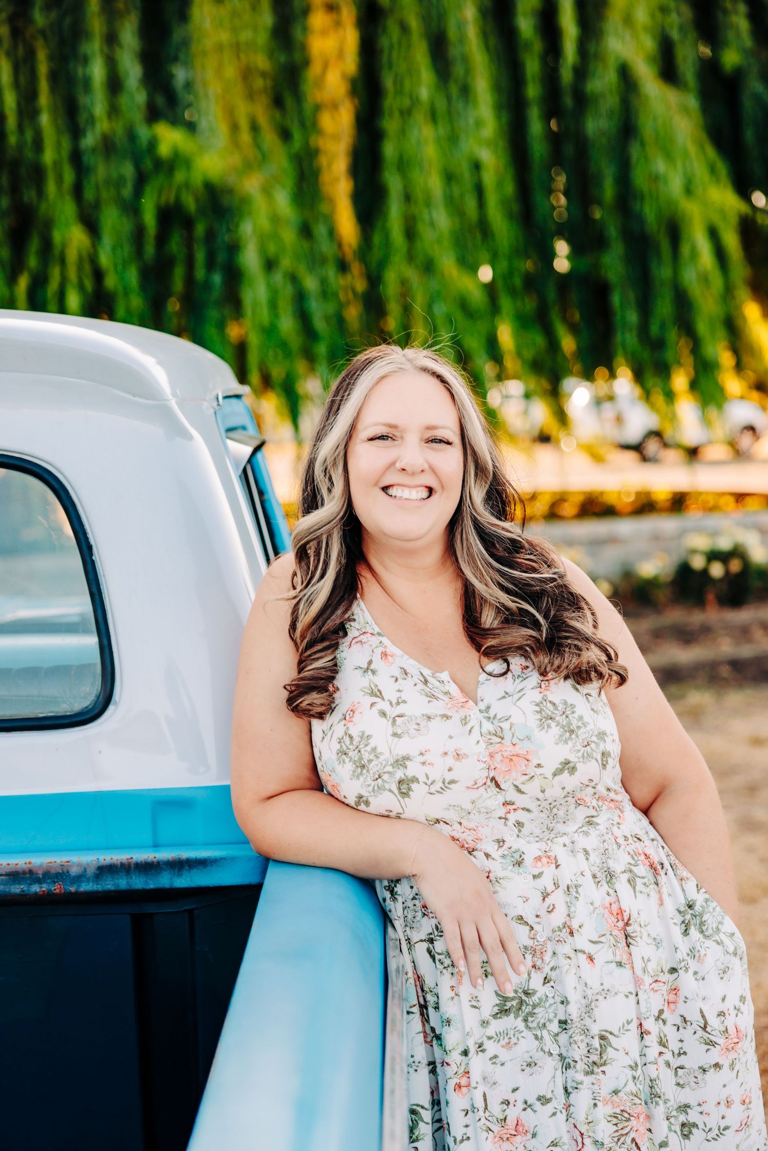 A woman in a floral dress poses happily against a classic blue car with willow trees in the background.