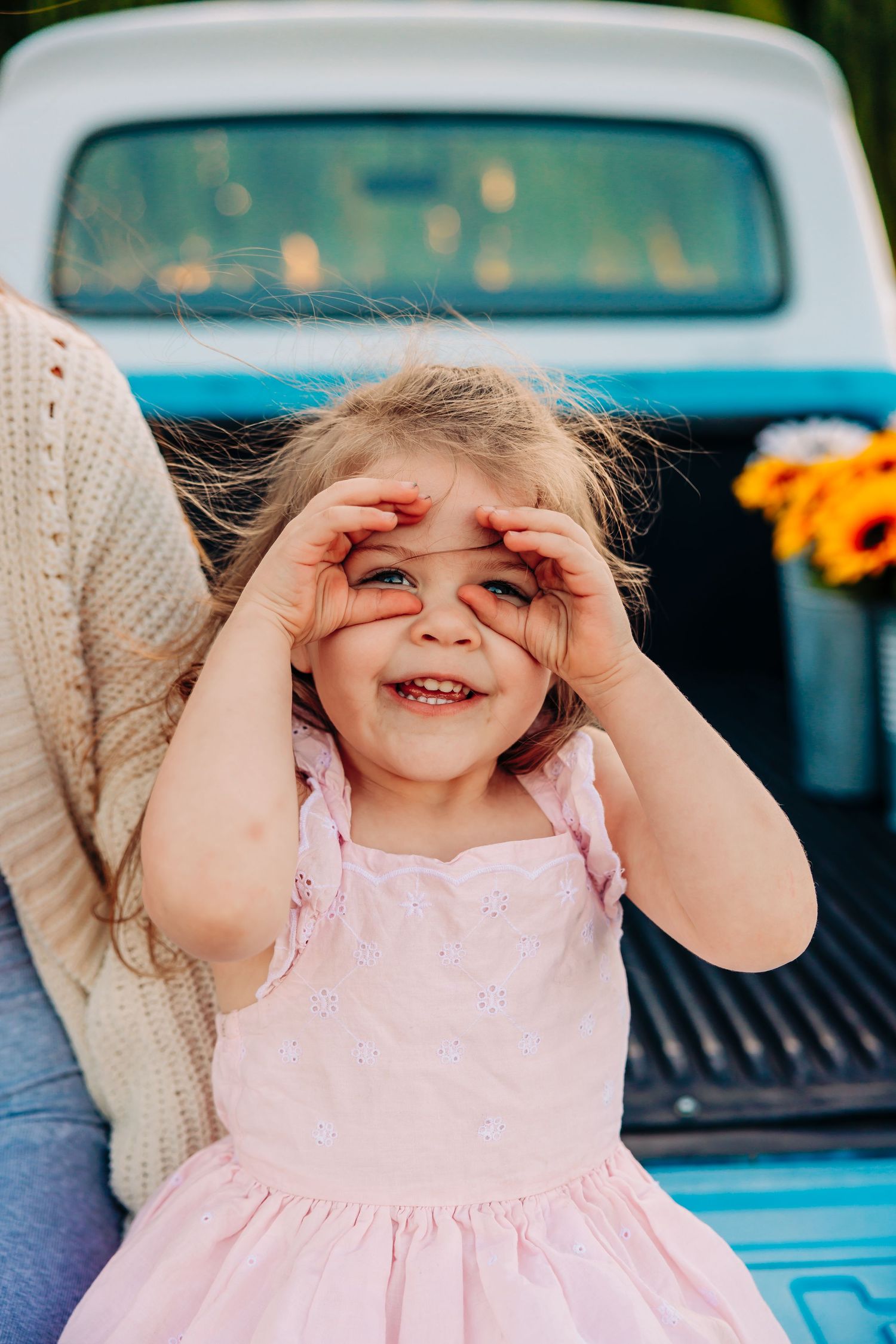 A child in a pink dress plays peek-a-boo while standing in front of a vintage blue truck.