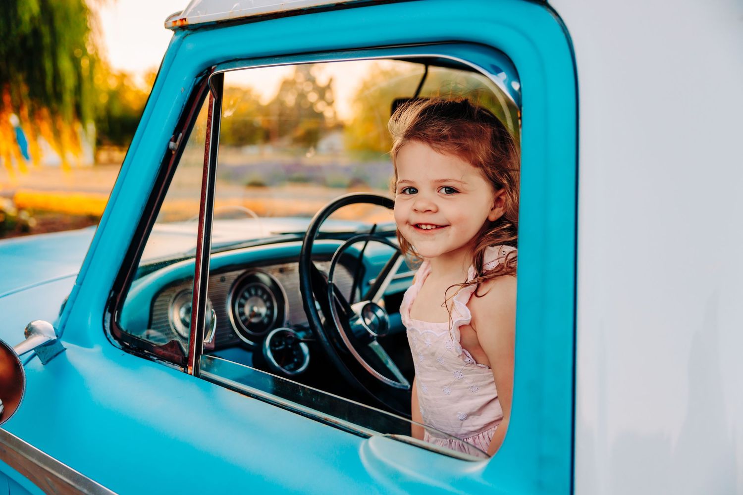 Young child sitting in a bright turquoise vintage car looking out the window during golden hour lighting.
