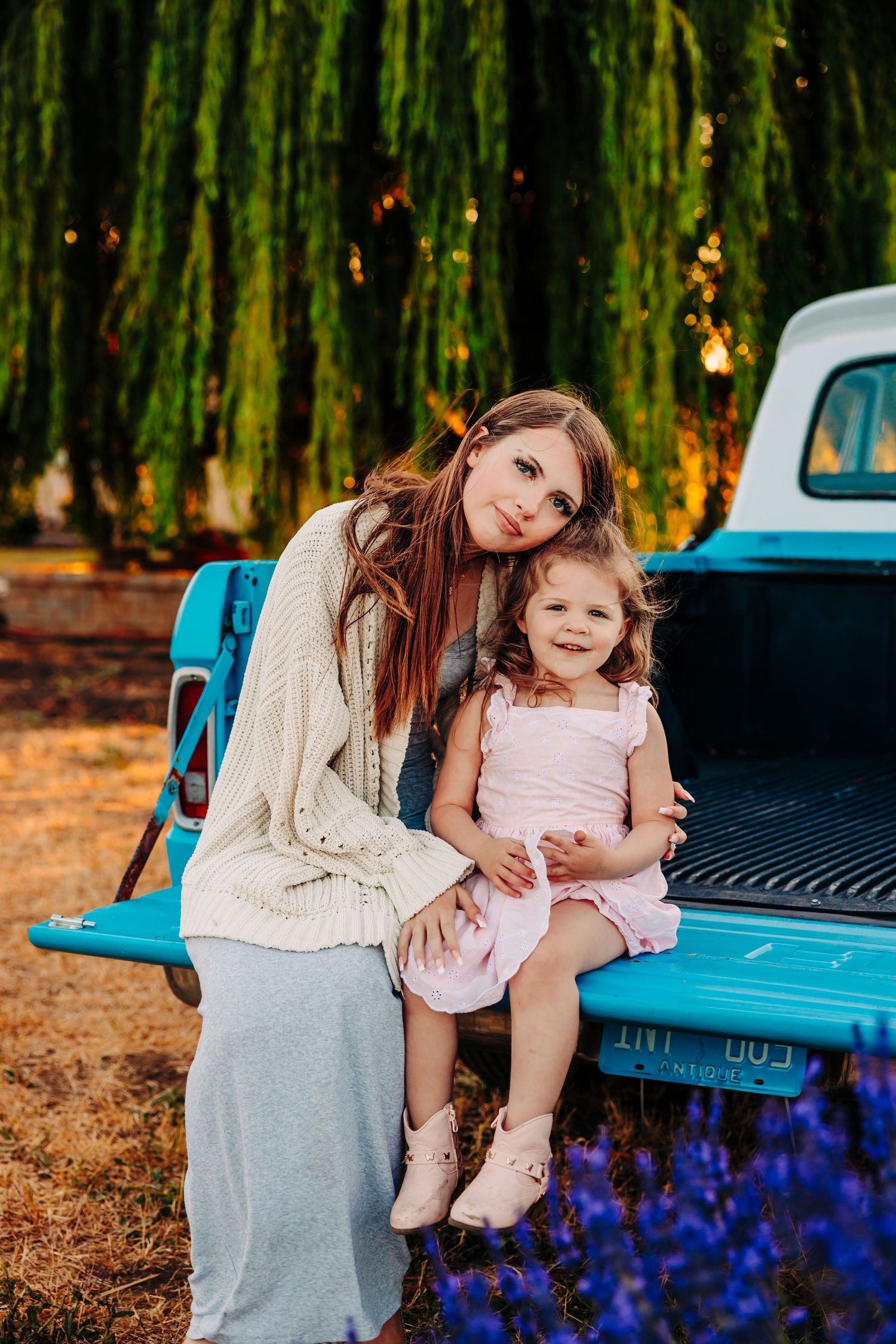 Mother and daughter share tender moments together by a vintage blue pickup truck surrounded by willow trees and flowers.