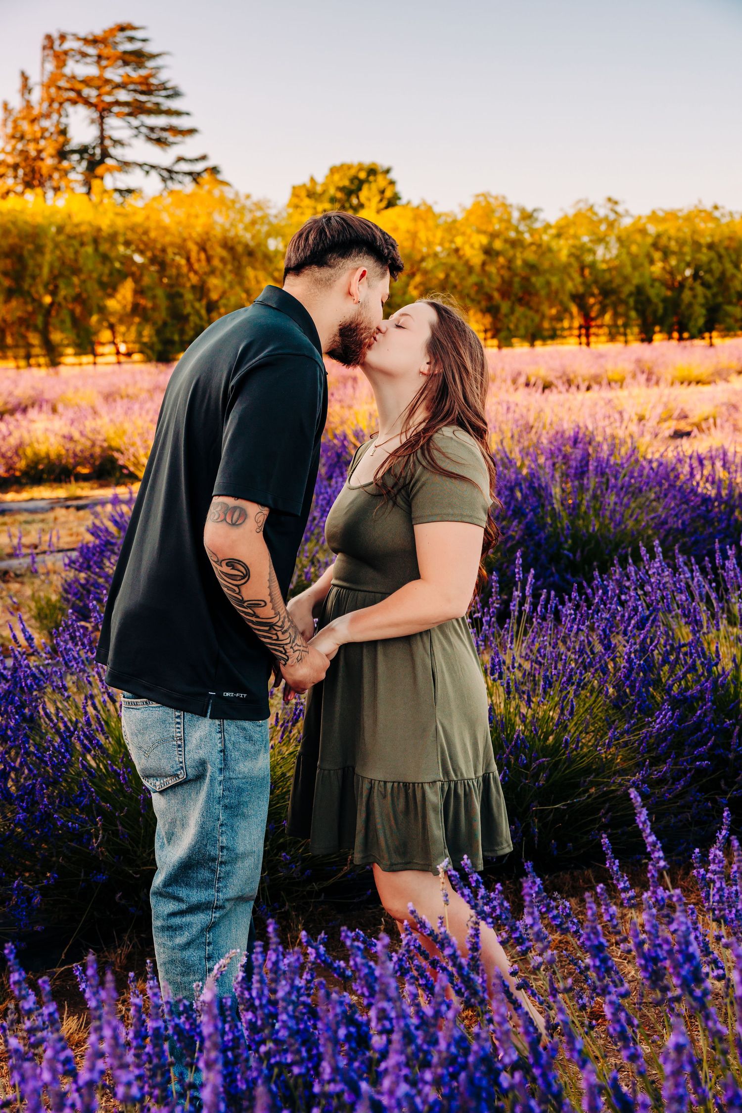 A romantic couple shares a kiss in a vibrant lavender field at sunset with golden trees in the background.