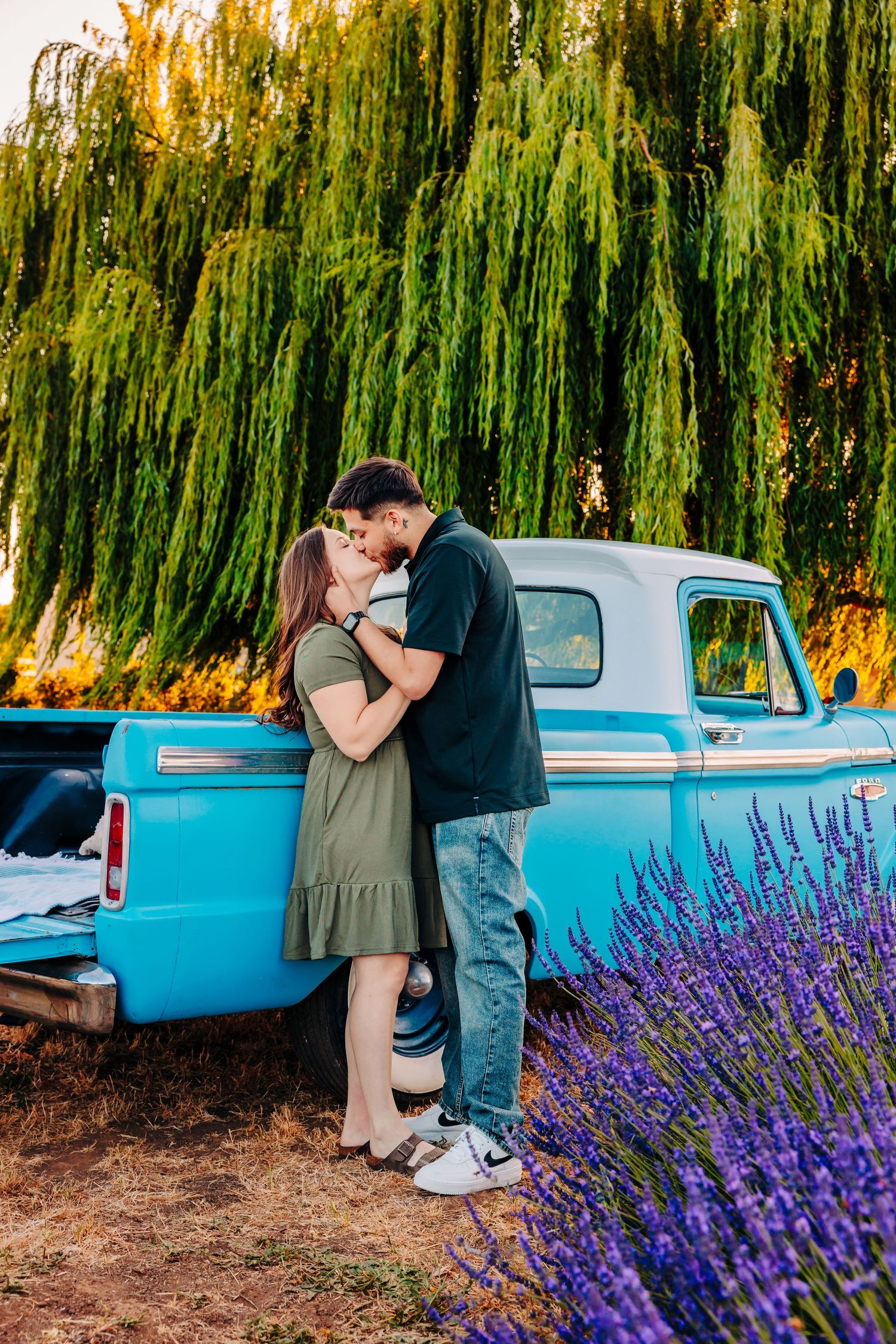 A couple embraces next to a vintage blue pickup truck surrounded by lavender and weeping willows.