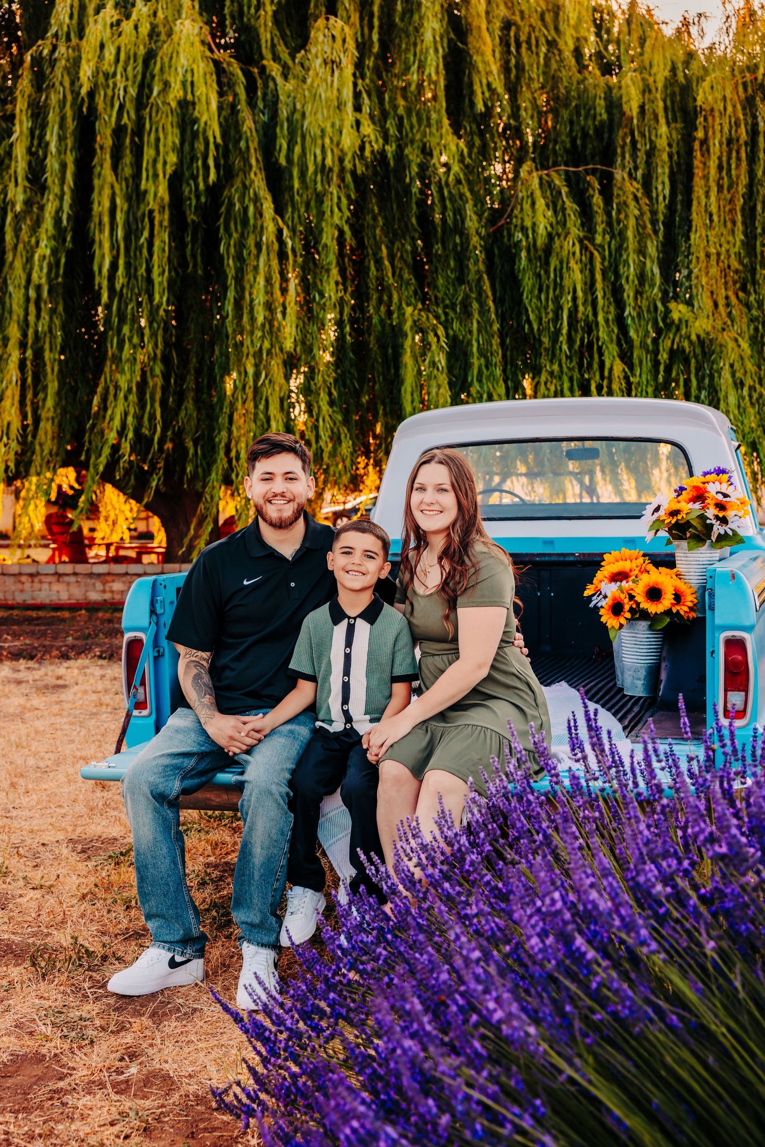 A family poses by a vintage blue pickup truck decorated with sunflowers amid lavender fields and willow trees.