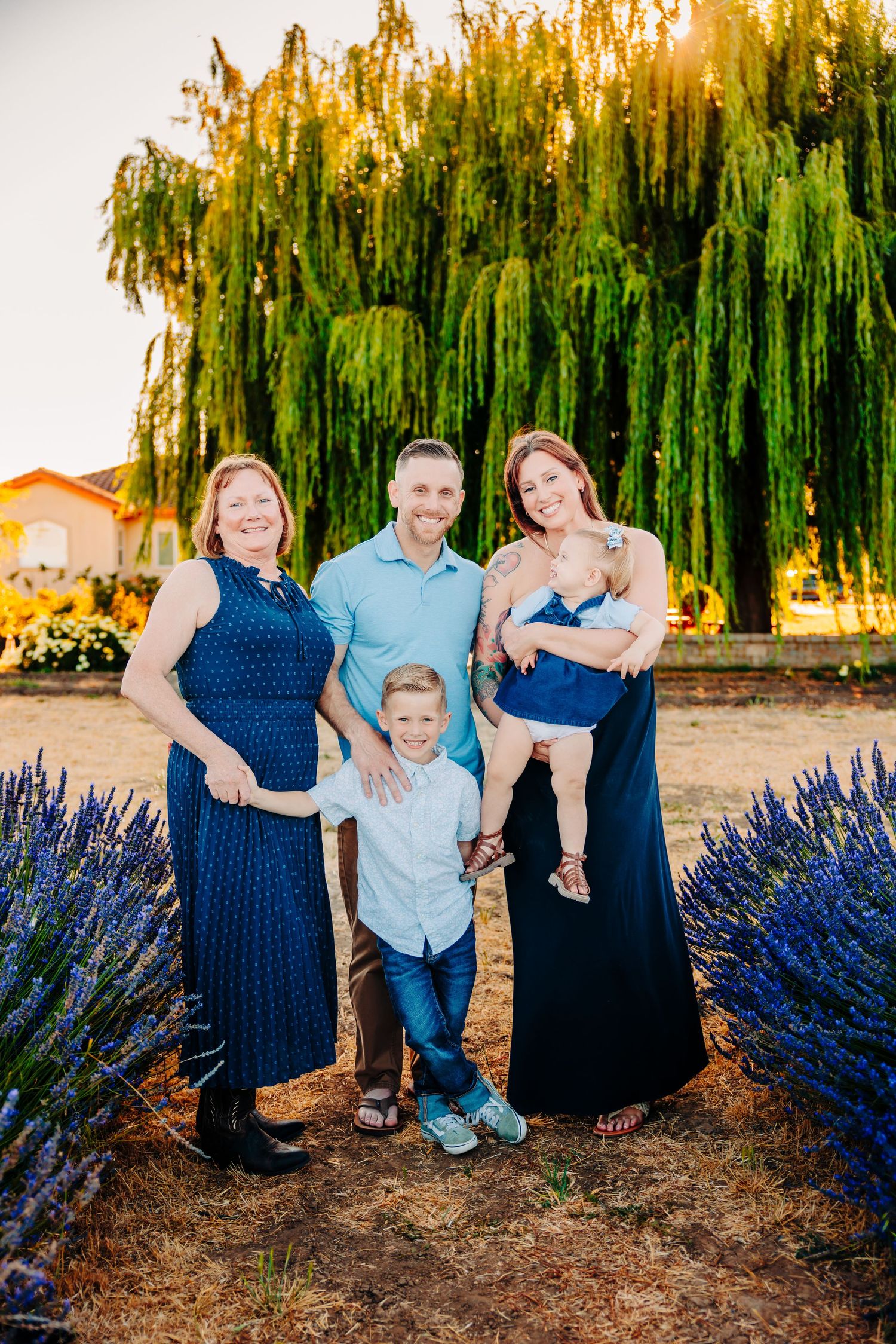 A family poses together in front of a weeping willow tree with vibrant purple lavender bushes at sunset.