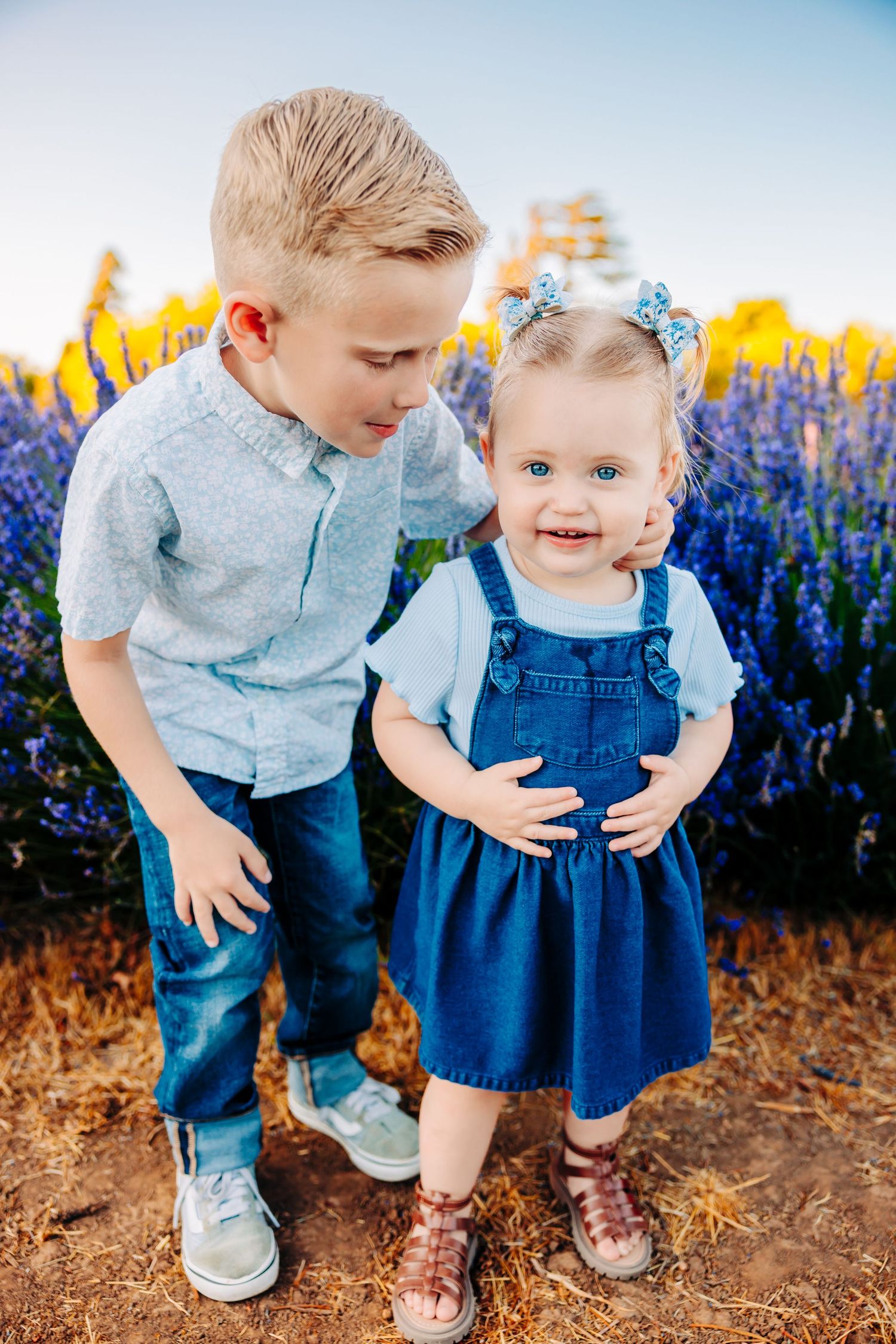 Two children in denim and blue clothing stand together among vibrant wildflowers on a sunny day.