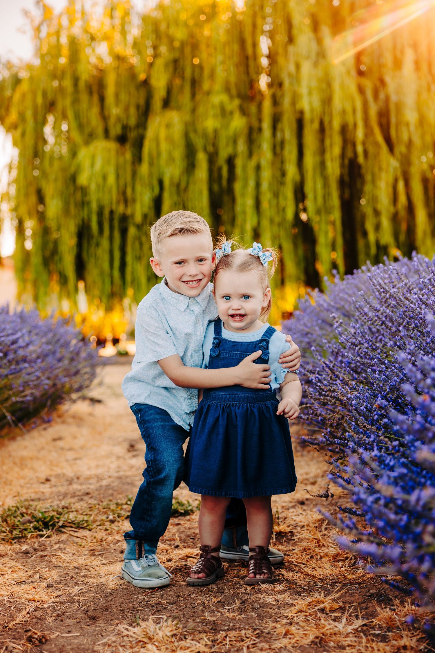 Two children pose together near blooming purple lavender flowers in a garden at golden hour.