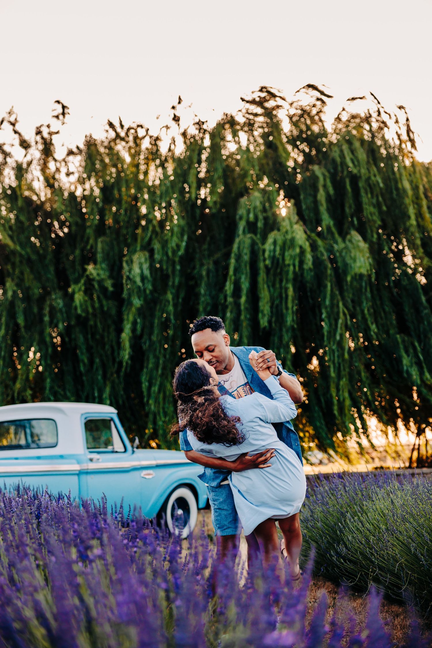 A romantic couple dances and embraces in a lavender field at sunset next to a vintage light blue car.