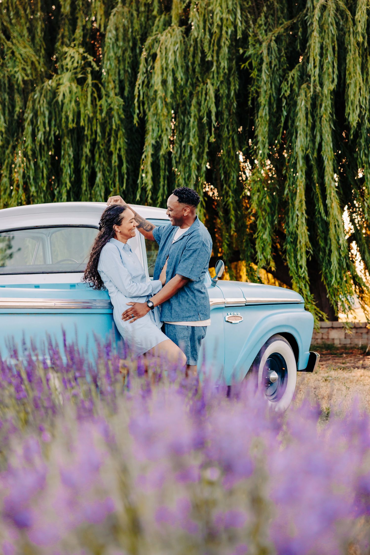 A couple shares a romantic moment beside a vintage light blue car with weeping willows and purple lavender flowers in view.