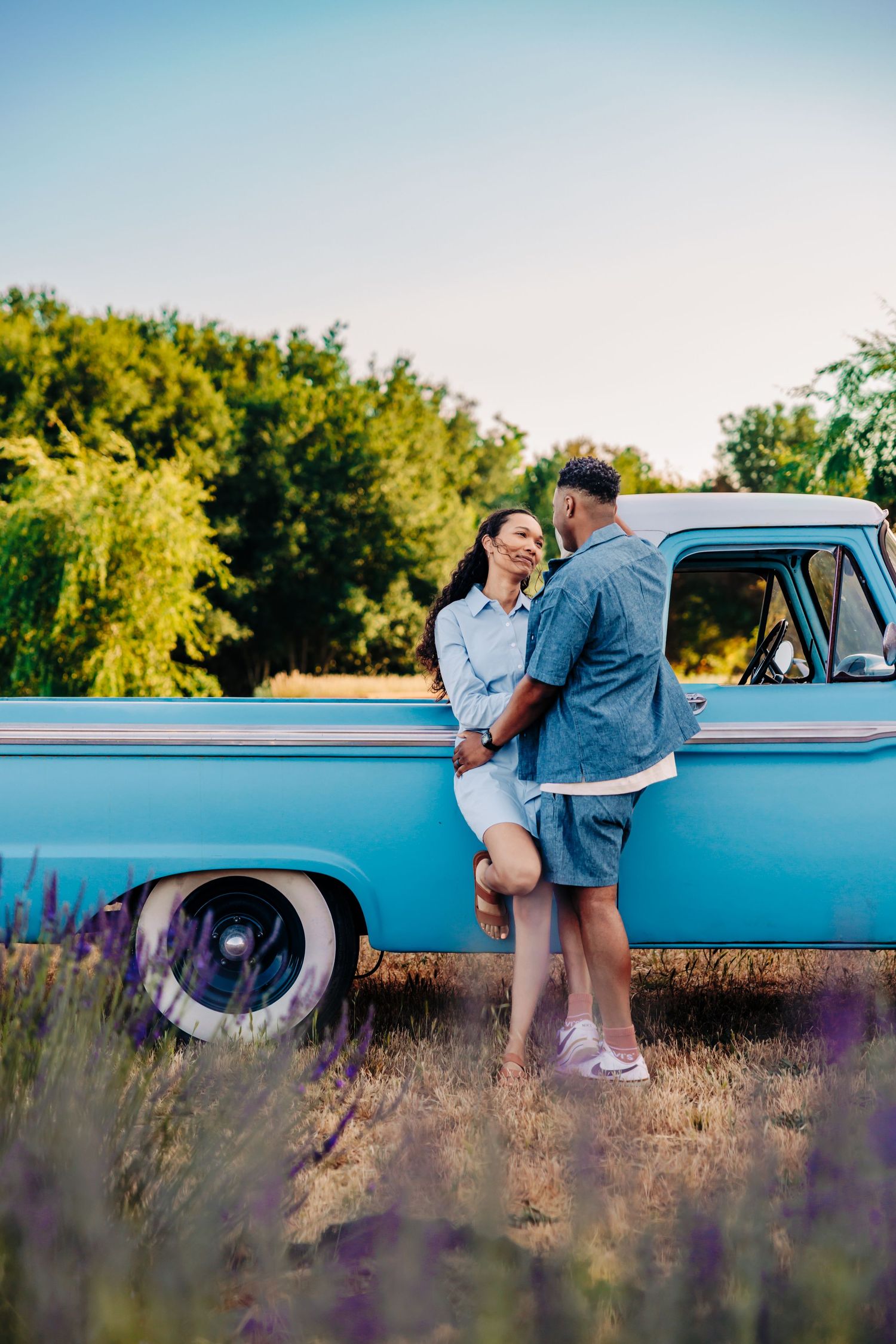 A couple embraces by a classic blue pickup truck in a field of blooming lavender on a sunny summer day.