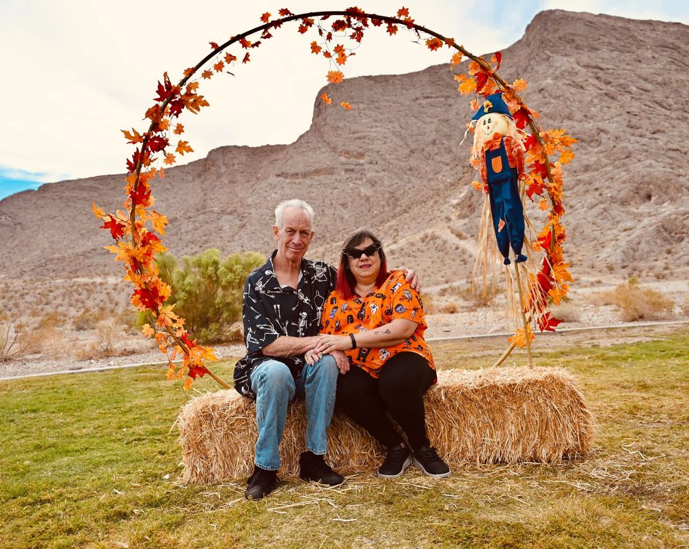 Couple sitting together on hay bales under a fall foliage arch with a scarecrow in a desert setting.