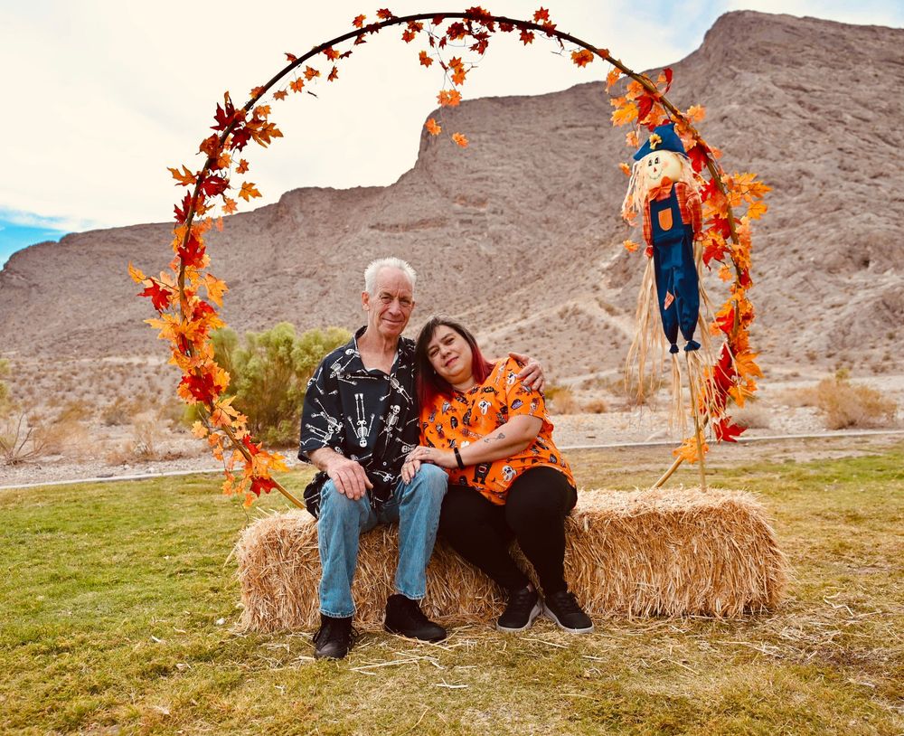 Couple cuddling on hay bales under a circular fall leaf arch with scarecrow in an outdoor desert scene.