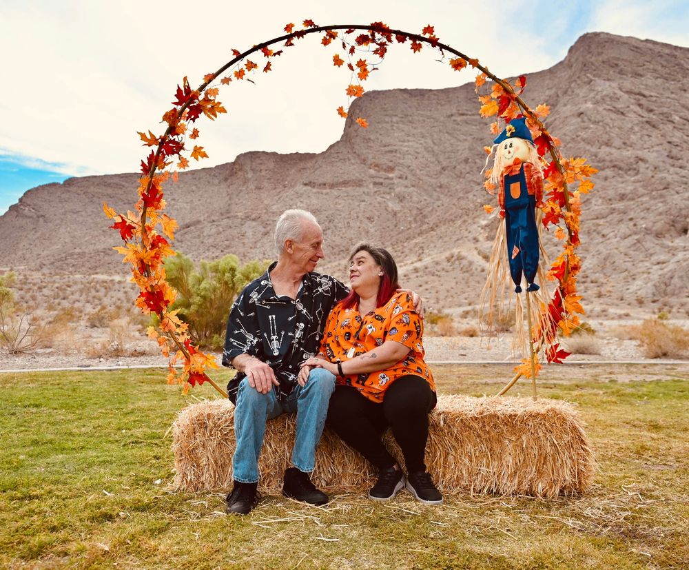 Couple sitting on hay bales smiling at each other under an autumn wreath arch in a desert landscape.