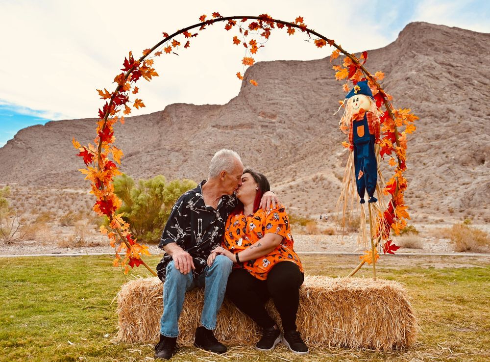 Couple sharing a kiss while seated on hay bales under a colorful autumn arch in a desert setting.