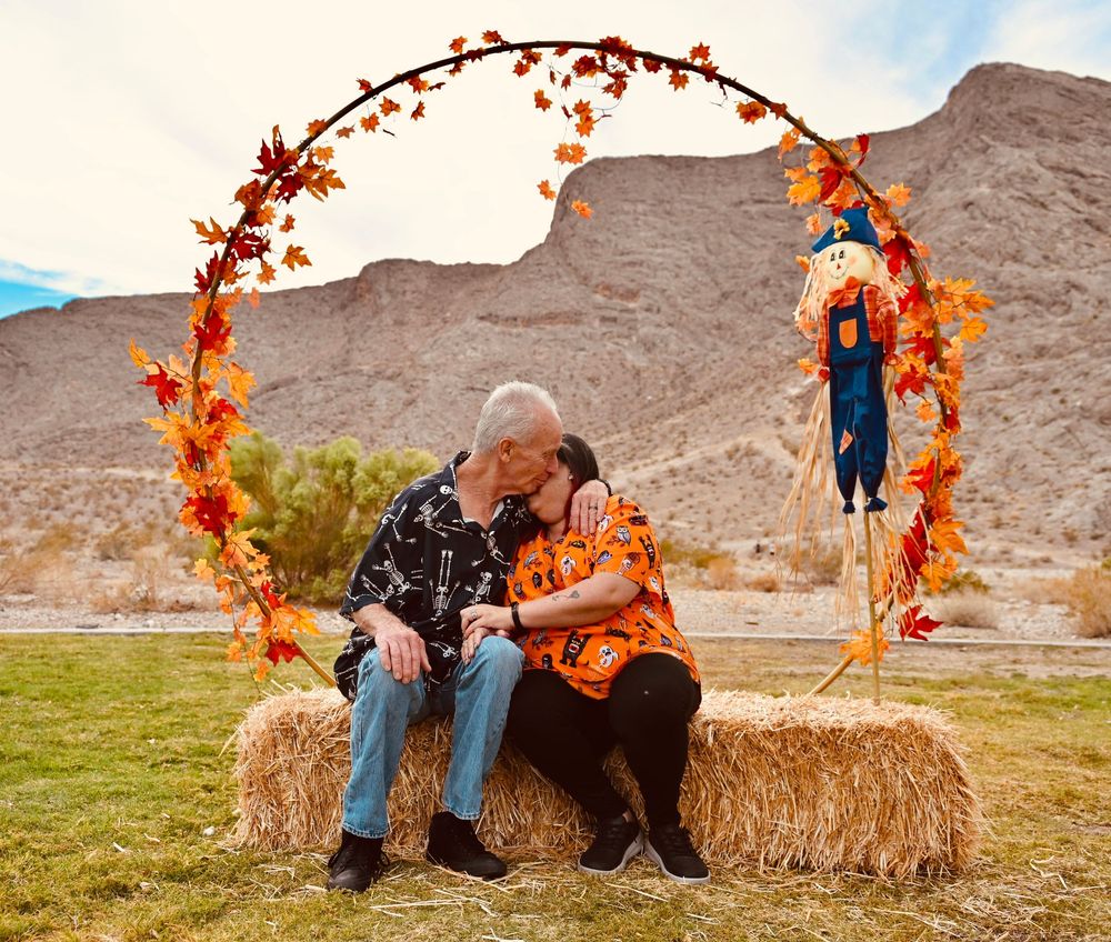 Elderly couple kissing on hay bale under fall leaf arch with scarecrow and desert mountains backdrop.