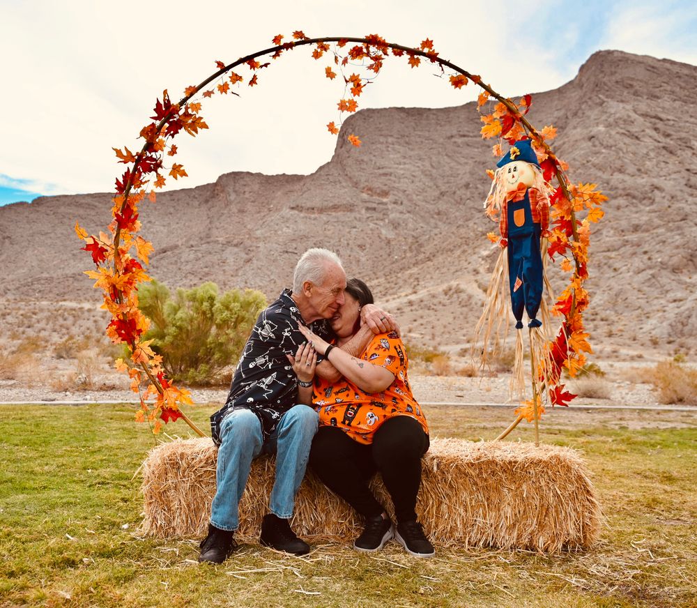Older couple embracing on hay bale beneath circular fall foliage arch with mountain scenery.