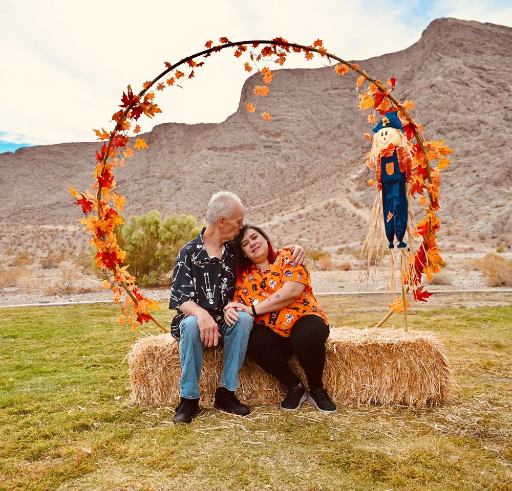 Senior couple sitting together on hay bale under decorative autumn arch in desert setting.