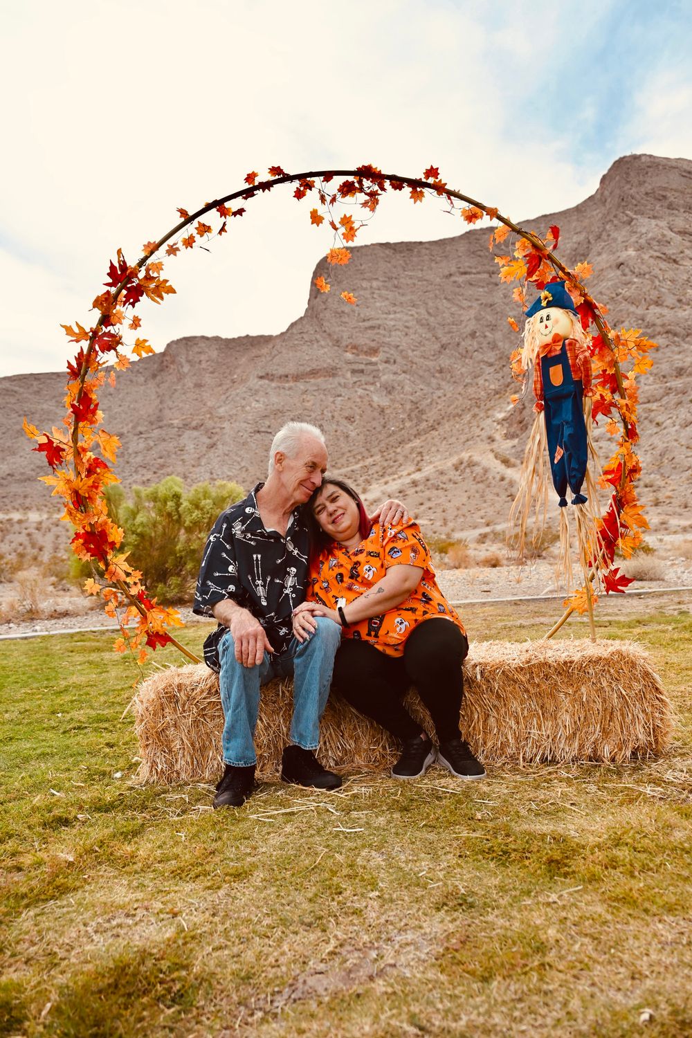 Couple sitting on hay bales embracing under a fall leaf arch with desert mountains behind.