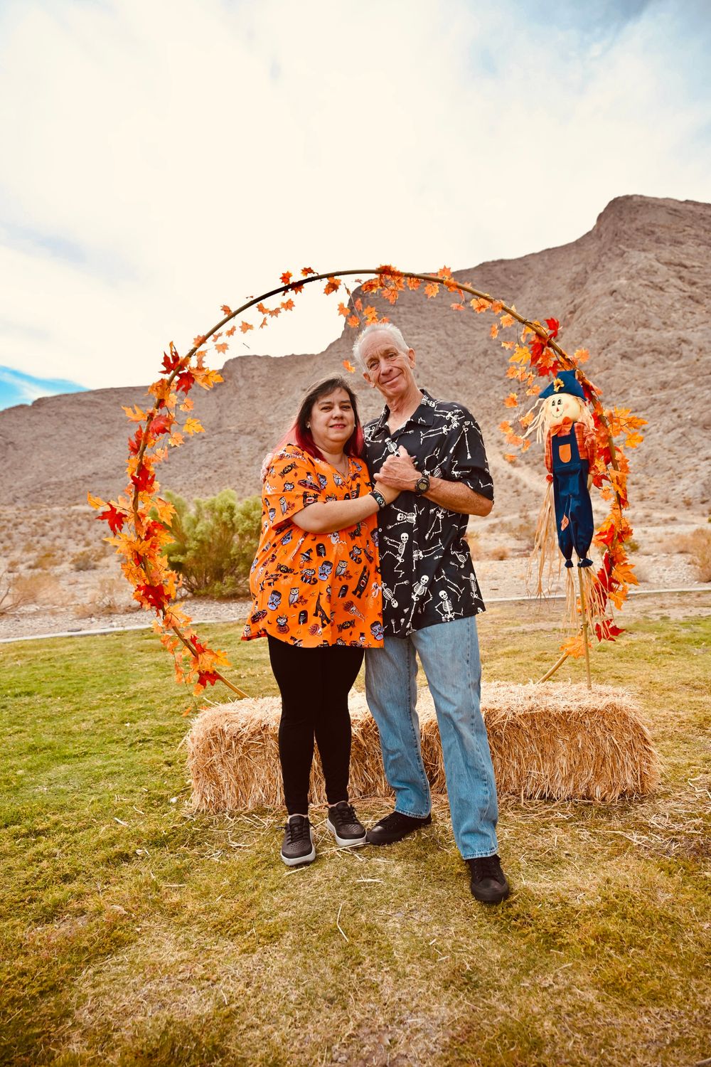 Couple in Halloween-themed shirts posing together under a fall leaf arch with scarecrow decor.