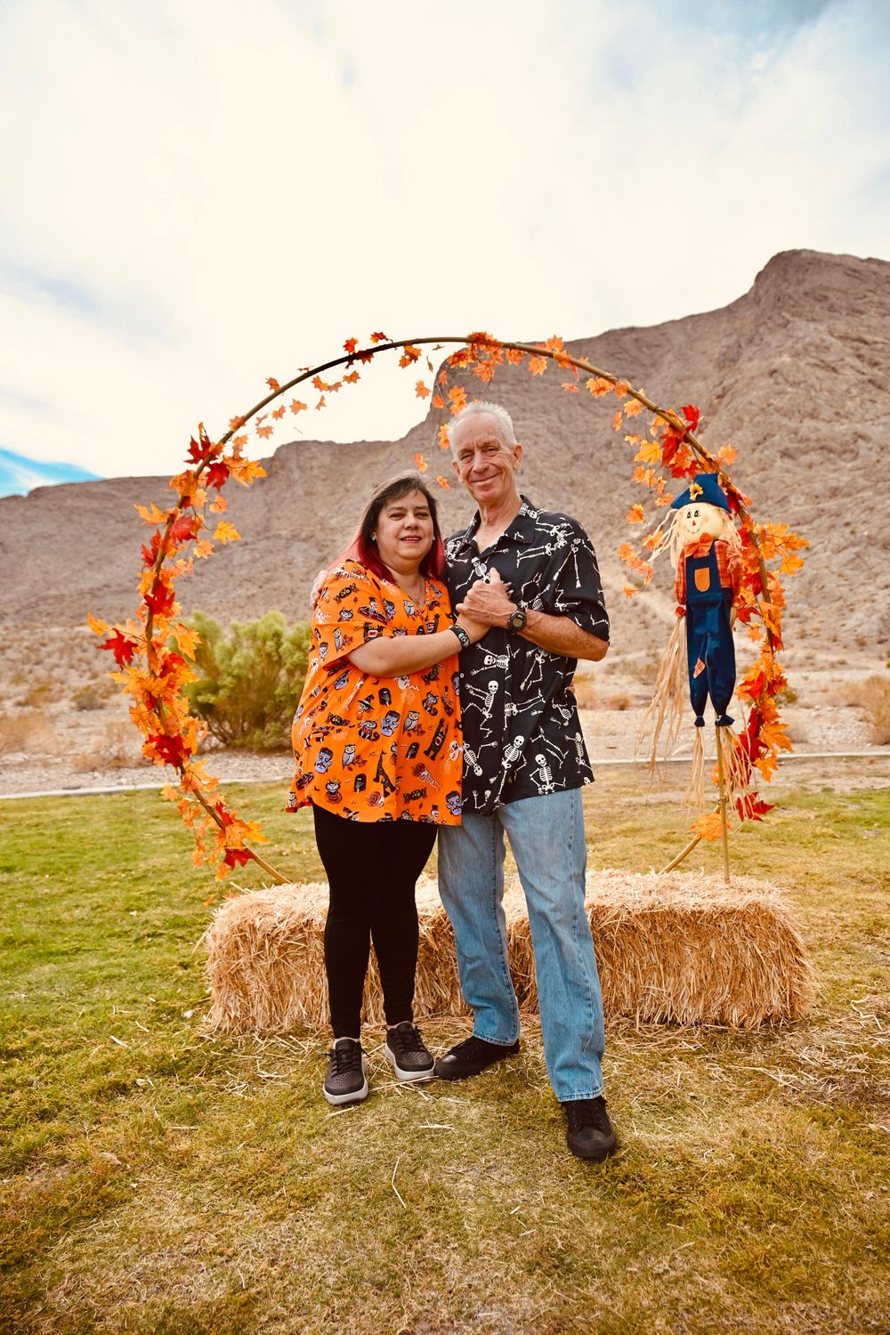 Smiling couple in Halloween shirts standing on hay bales under an autumn wreath arch outdoors.