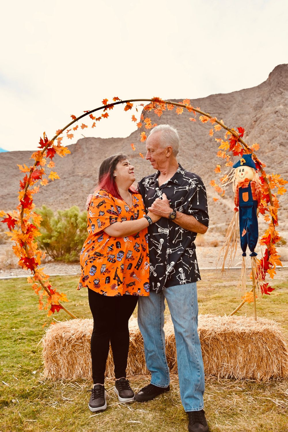 Couple in Halloween attire dancing and laughing under an autumn leaf arch with scarecrow nearby.