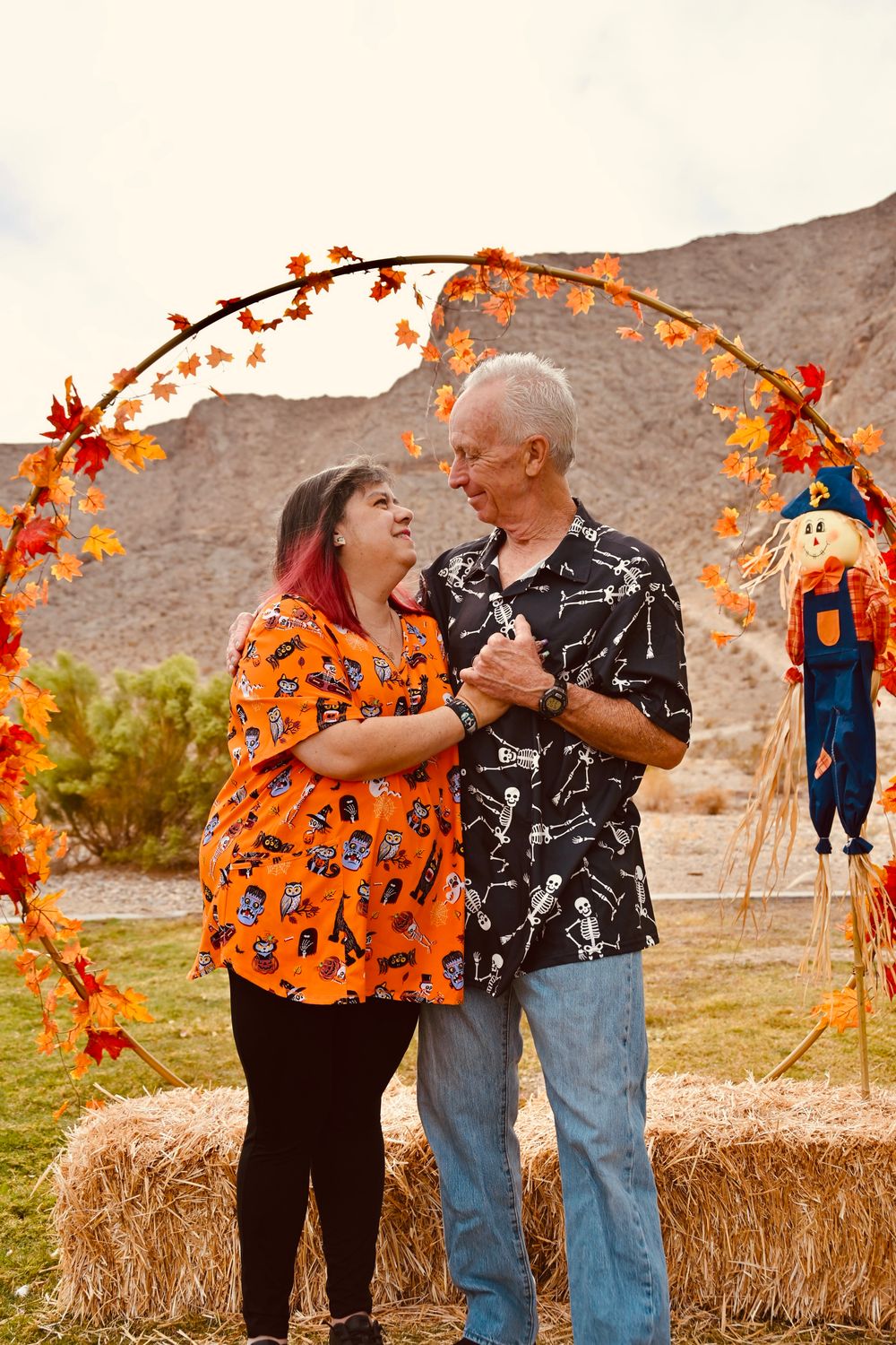 Couple dancing and smiling under a fall leaf arch with hay bales and scarecrow in a desert setting.