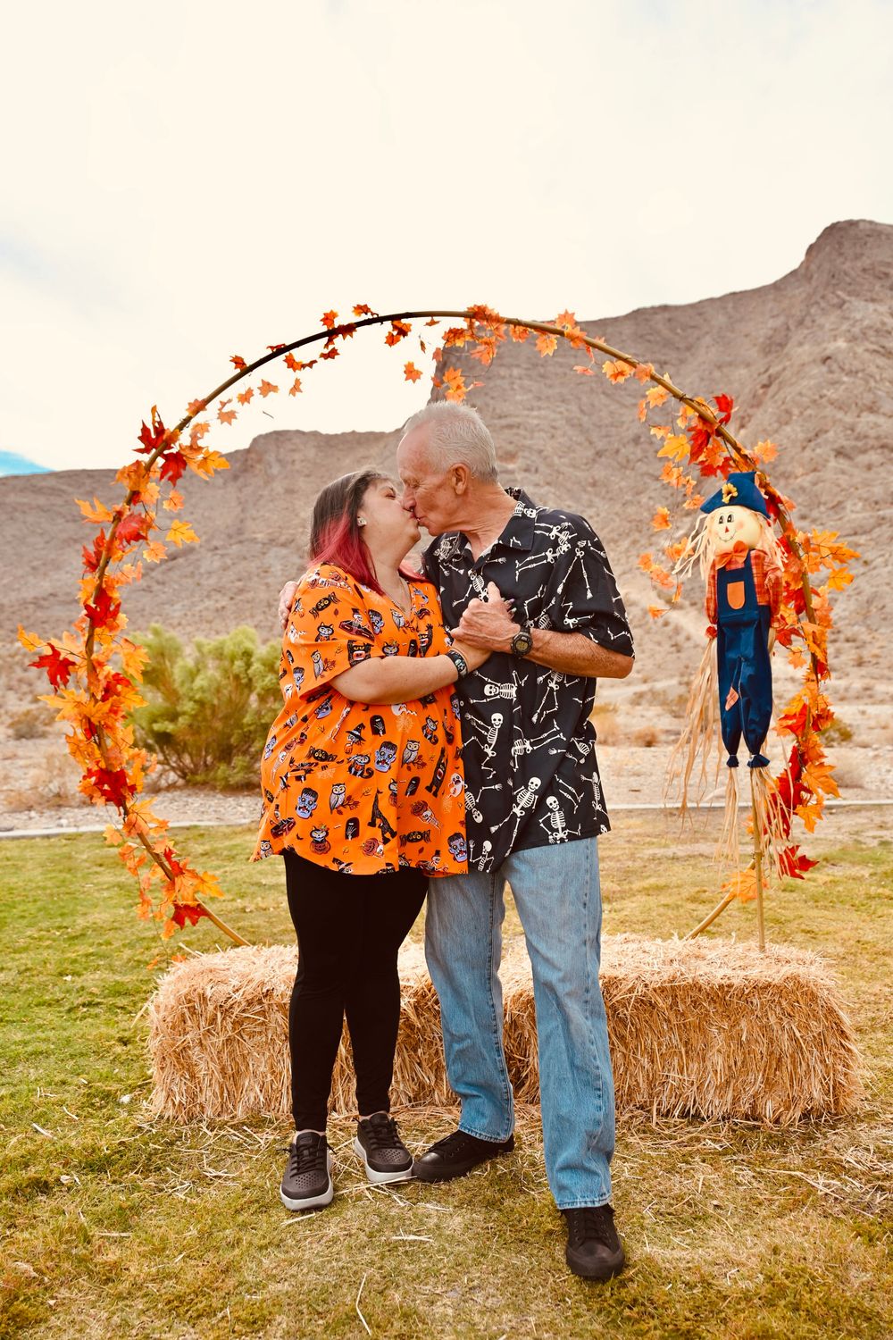 Couple sharing a kiss under an autumn wreath arch with hay bales and scarecrow in a desert landscape.