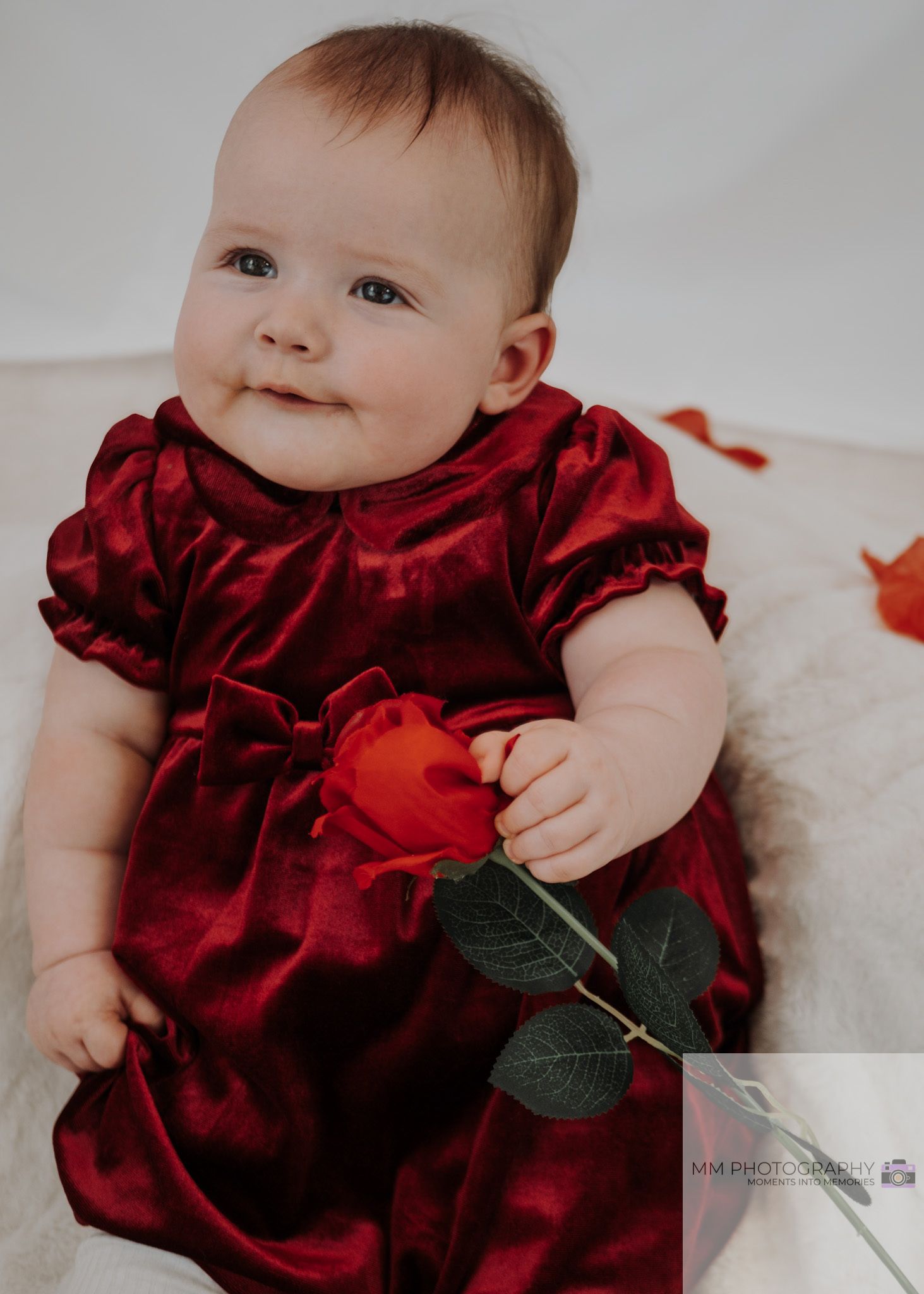 A baby in a deep red velvet holiday dress sits on a white surface with festive decorations.