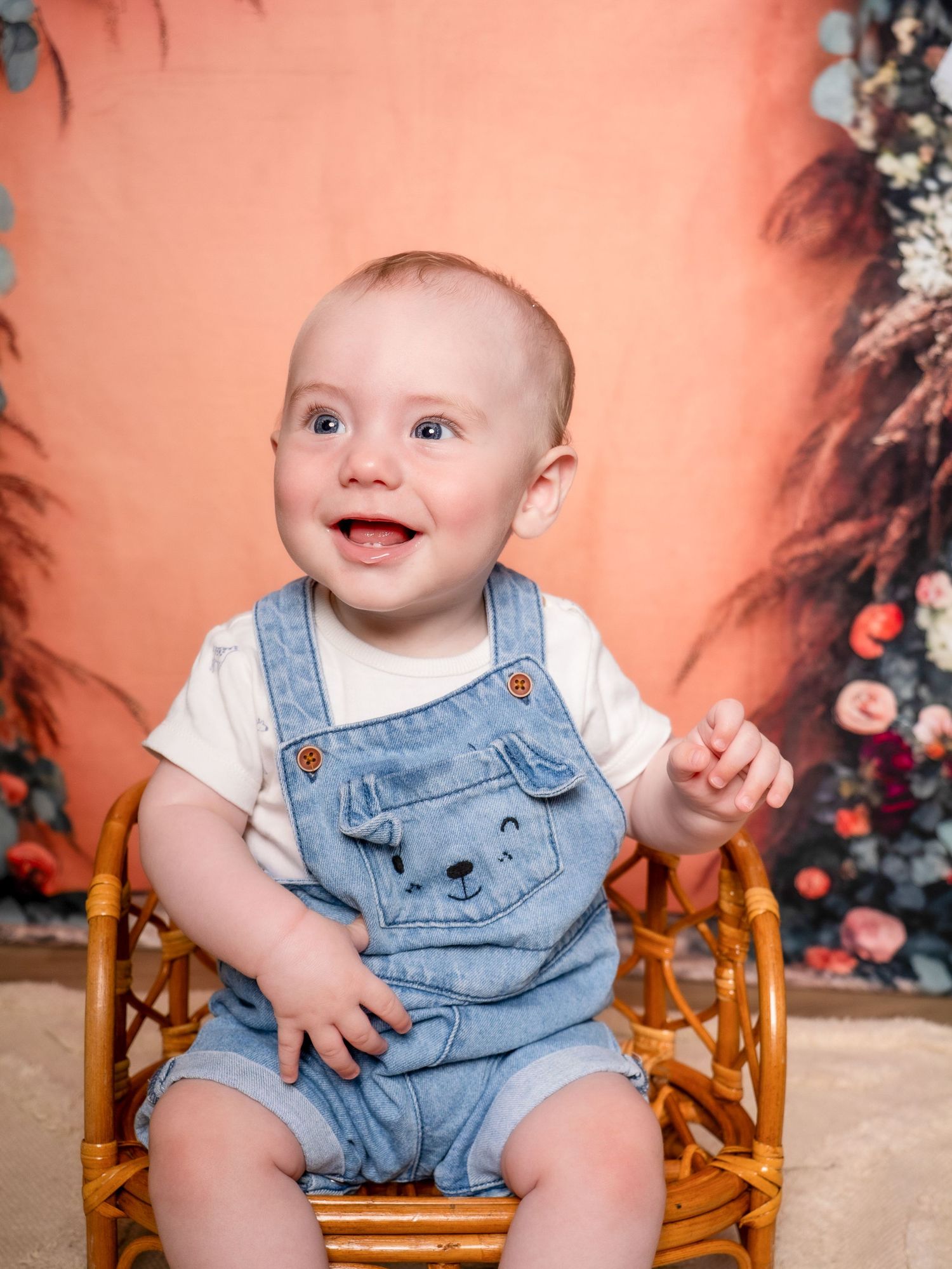 Baby wearing denim overalls and white shirt sits in wooden rocking chair against peach-colored backdrop with floral decor.