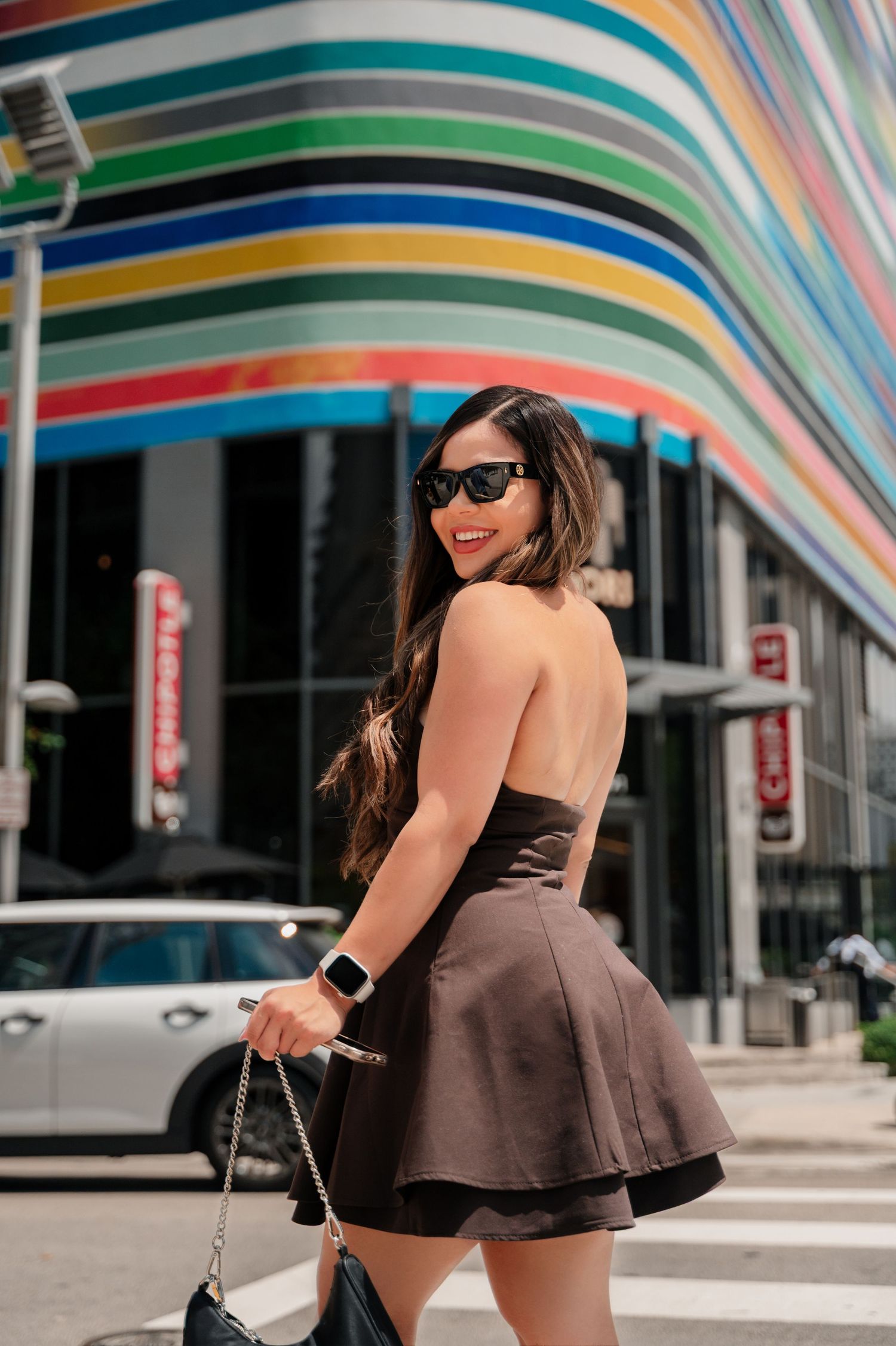 Stylish woman in brown romper and sunglasses poses playfully in front of colorful striped wall art installation.
