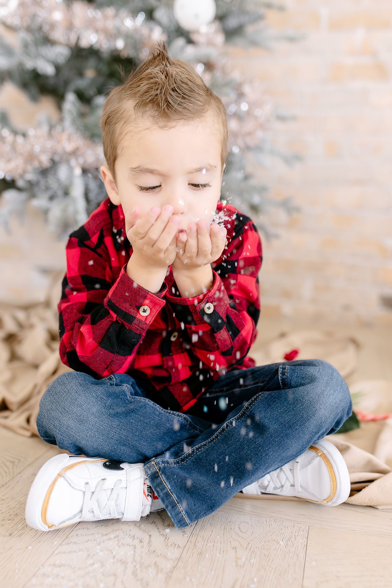 A child in a red plaid shirt and jeans sits cross-legged and blows glitter in a festive holiday photoshoot setting.