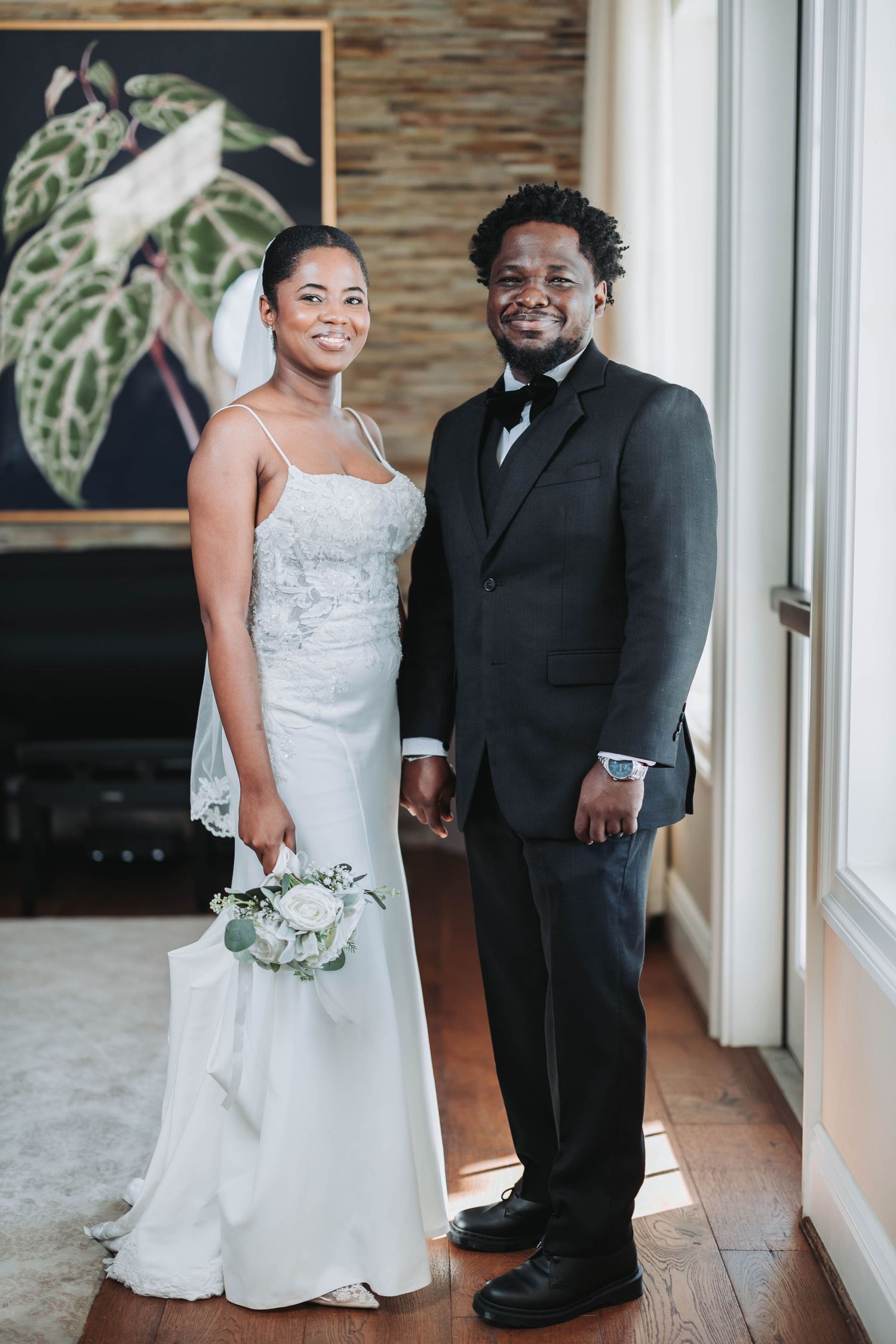 A bride in a white spaghetti strap gown and groom in black tuxedo pose together in an elegant indoor setting.