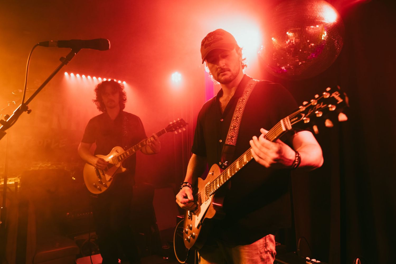Two guitarists performing on stage bathed in warm red and orange stage lights at a live concert.
