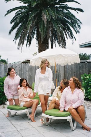 Group of friends in pink and white loungewear relax outdoors on patio furniture under palm trees.