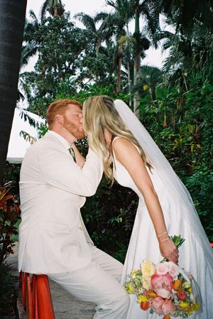 A couple in white wedding attire share a romantic kiss in a tropical garden setting.