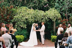 Outdoor garden wedding ceremony with guests seated among lush greenery and flowering trees.