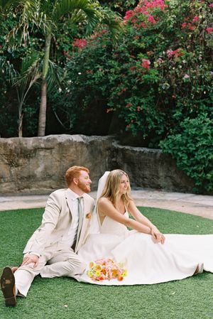 A couple in white attire relaxes on green grass with pink flowering bushes in the background at a garden venue.