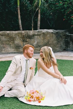 Newlyweds relax on grass with colorful bouquet in tropical garden setting.