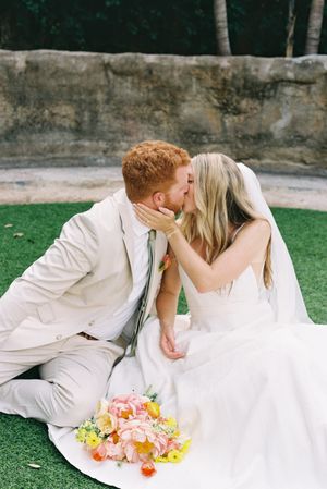 Newlyweds in white sharing a romantic moment on grass with colorful bouquet.