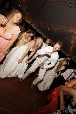 Wedding guests dance energetically on a wooden dance floor under string lights at an evening reception.