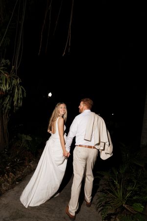 A couple in wedding attire walking away from the camera through a dark garden path at night.