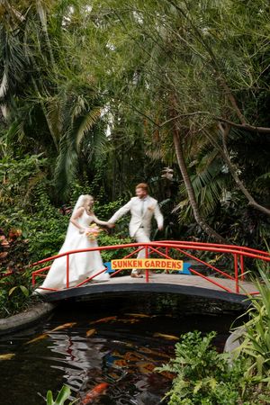 Couple crossing red Japanese-style bridge in garden setting wearing wedding attire.