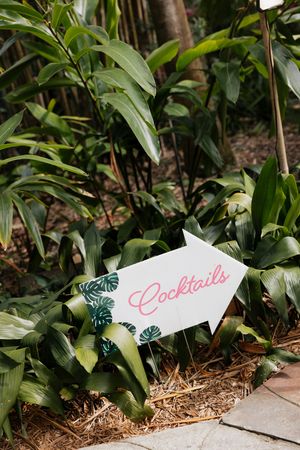 White arrow signs with pink text reading 'Cocktails' point the way through tropical foliage.