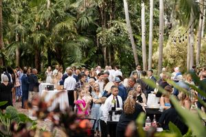 Wedding guests mingle outdoors among tropical palm trees and lush greenery at a garden reception.