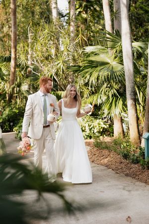 Wedding party walks along a palm tree-lined path in cream and white attire.