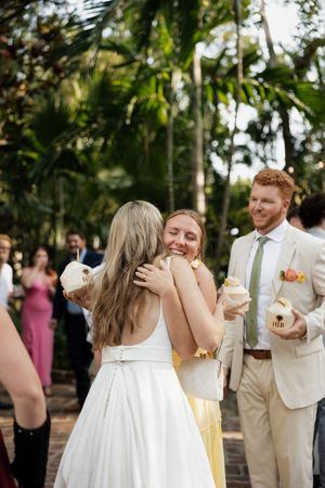 Wedding party members share emotional embraces during outdoor tropical reception with palm trees in background.