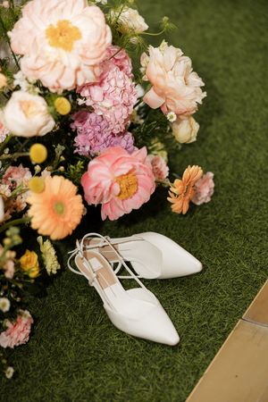 Elegant white bridal shoes placed beside a lush arrangement of pink and peach garden roses on grass.