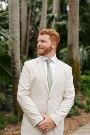 A person in a cream colored suit and sage green tie poses outdoors against a backdrop of tropical foliage.