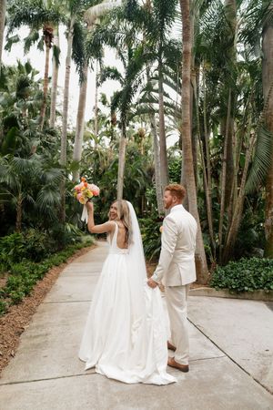 A wedding couple walks along a palm-lined pathway with the bride holding up a colorful bouquet.